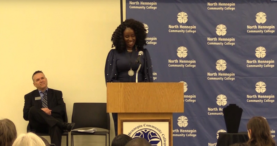 woman presenting on a stage in front of a podium man sitting behind her