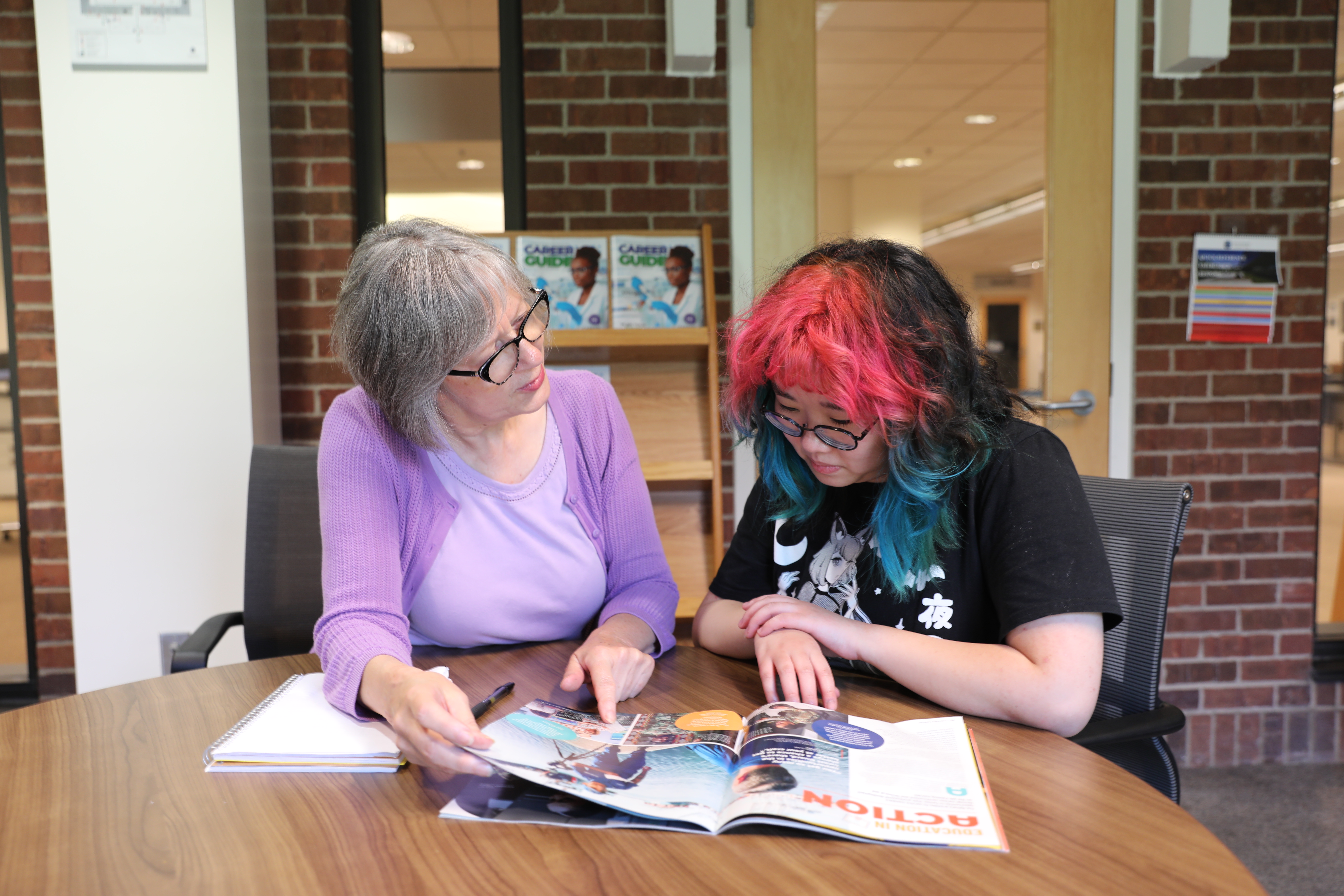 NHCC Workforce Innovation and Experiential Learning Center Director, Karen Philbin, sits with a female student at a table in the library looking at a career magazine together