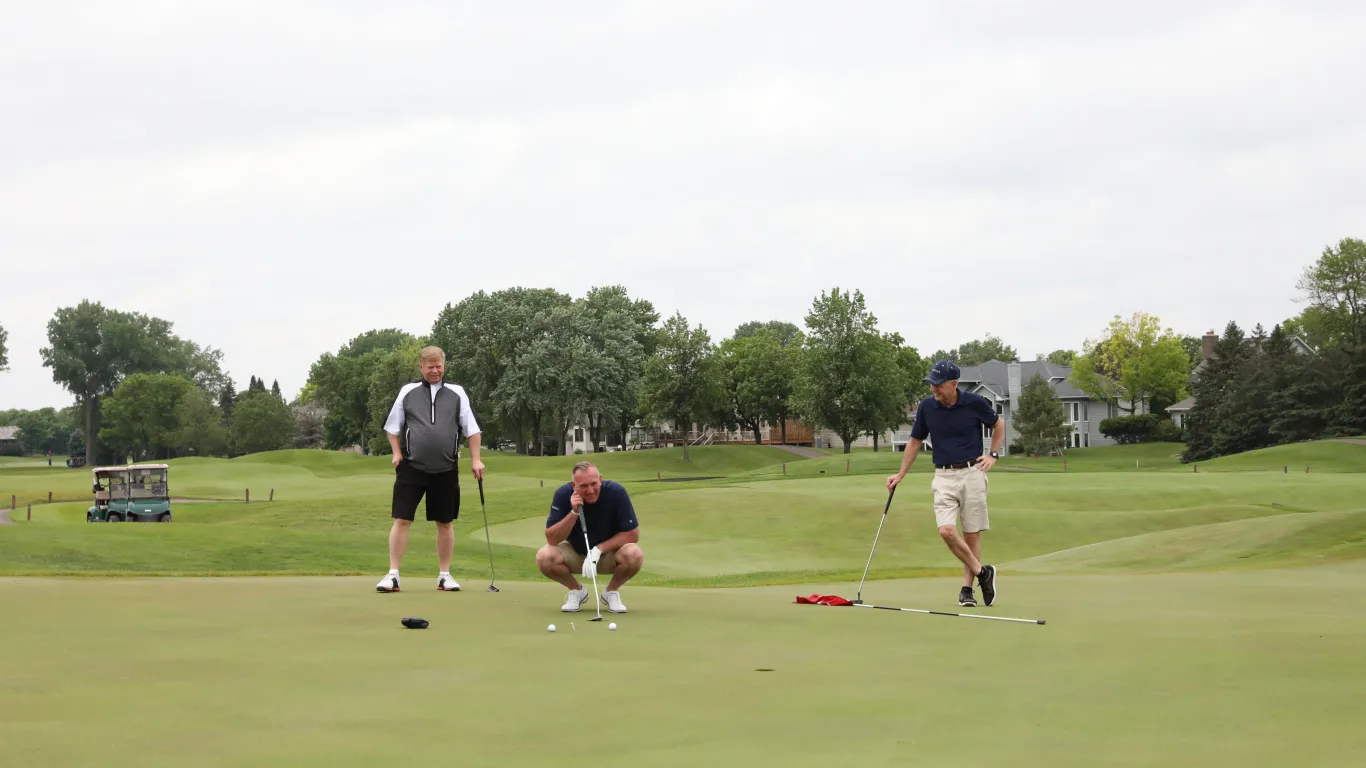 2 men watching a third man lining up to tee a golf ball into the hole