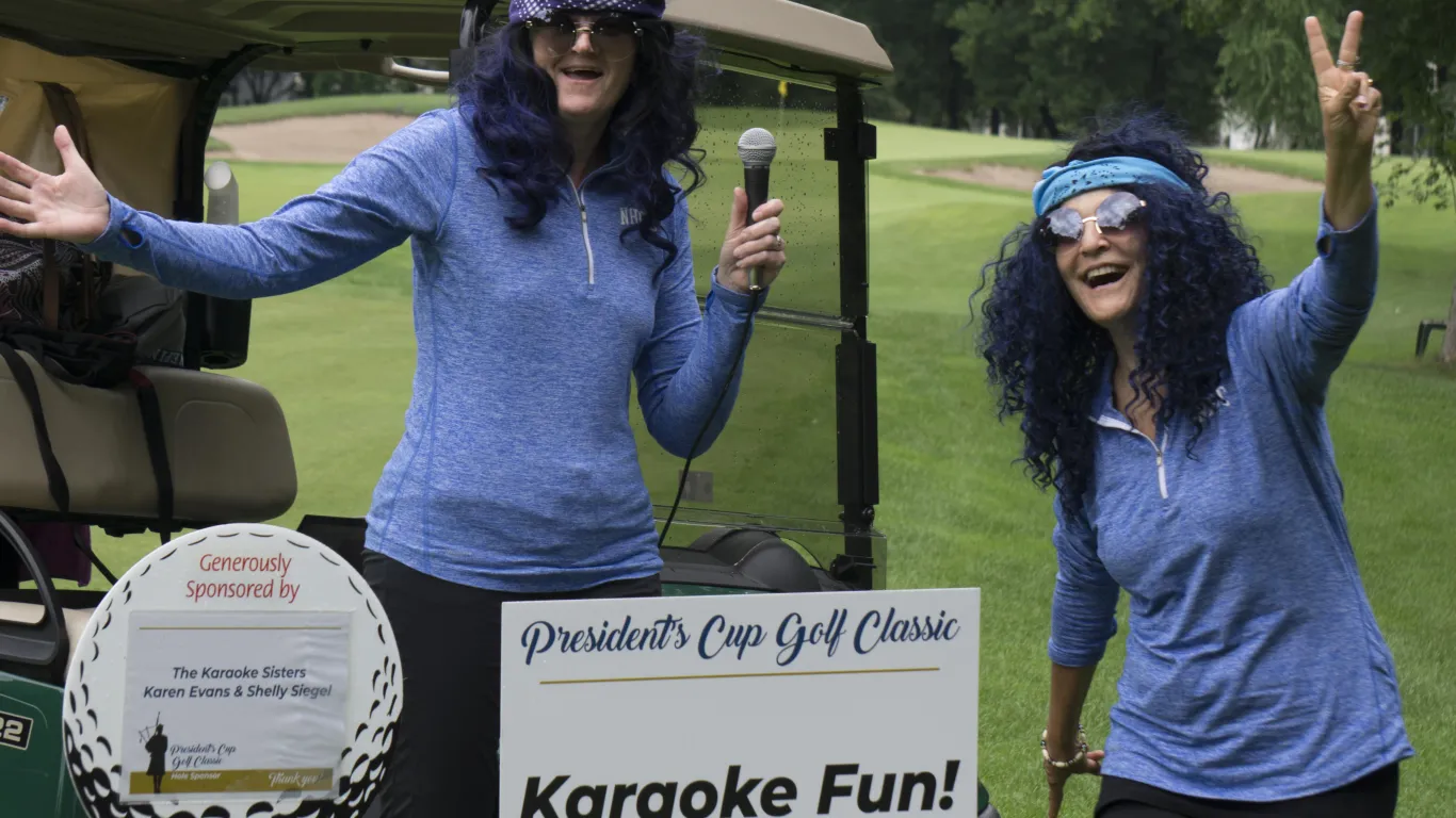 two women in wigs in fun poses in front gold cart and behind a sign reading karaoke fun