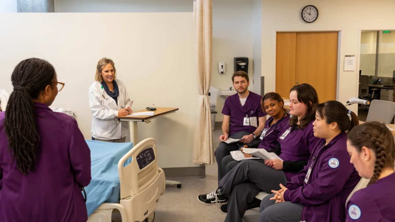 a nursing instructor speaking to a group of students in a nursing lab. 