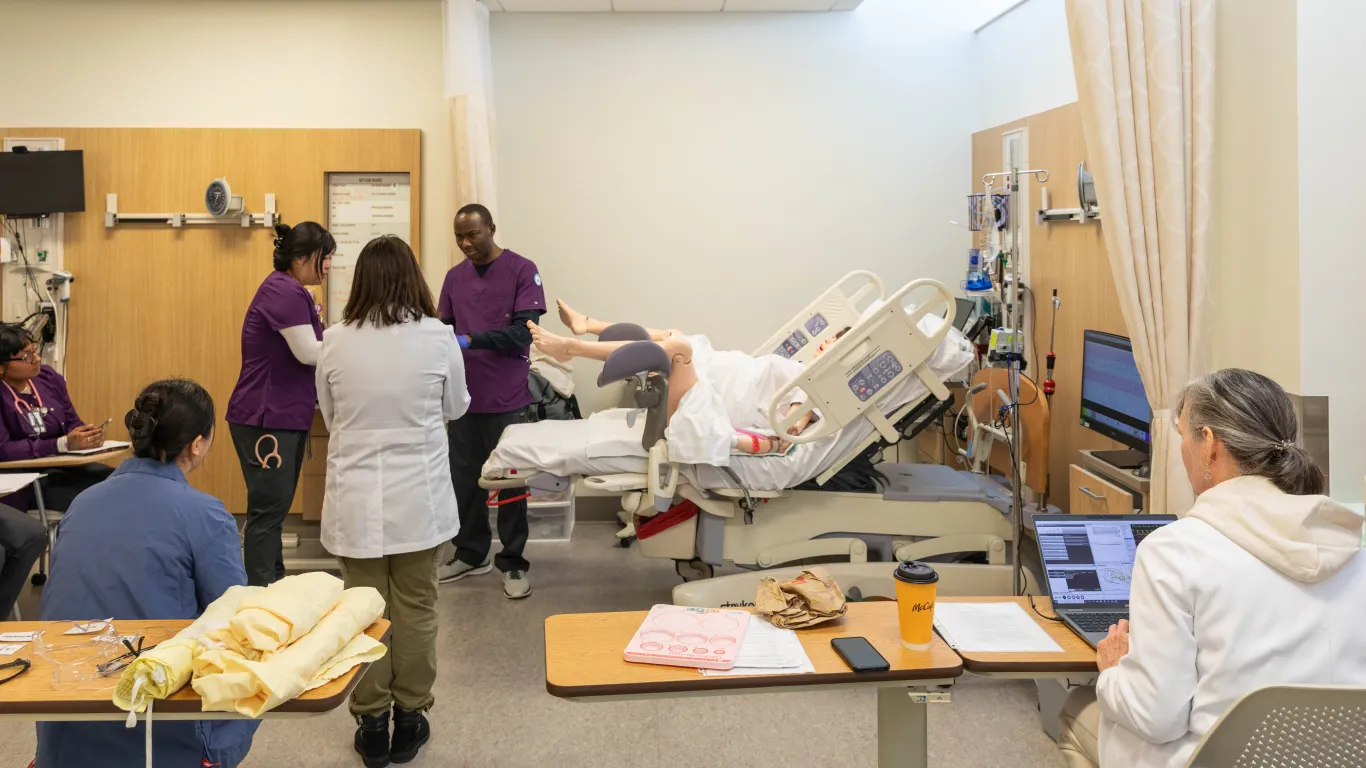 a group of students standing by a mannequin delivering baby with an instructor watching. 