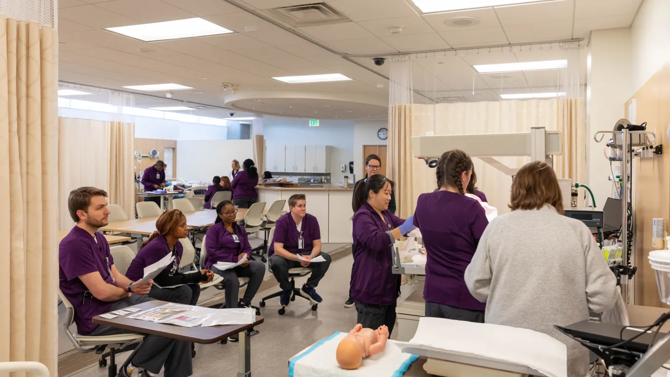 a group of students working by a table in the nursing lab with another group of students watching. 