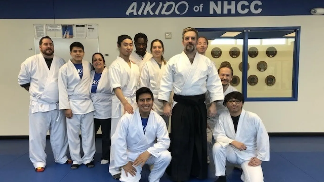 this is a group photo of NHCC instructor, Mark Larson standing with ten of his aikido students in the dojo on-campus, they are all wearing white robes and white pants, smiling for the photo. A sign in the background reads, 'Aikido at NHCC.' 