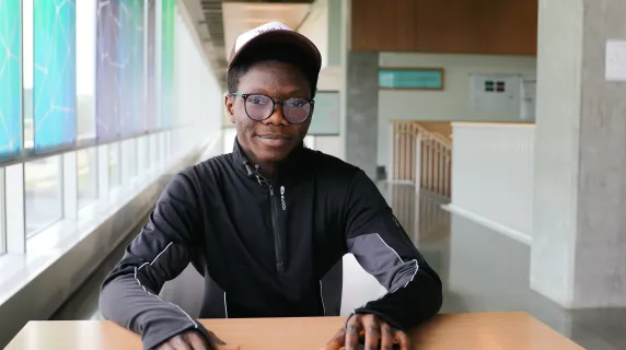 student with wearing a hat and glasses sitting at a table looking at the camera. 