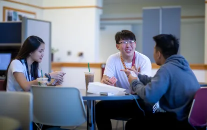 three students sitting at a table playing cards 