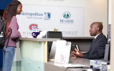 female student standing at university center talking with male faculty member sitting behind counter