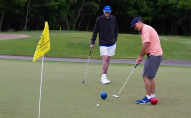 two men standing by a golf hole one about to put the ball into the hole