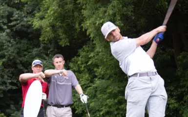 three men stand on a golf course, one hitting a golf ball