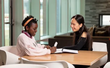 Two students sit at a table assisting each other with homework laid out on the table.
