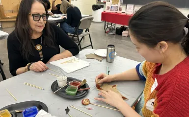 Artist Fern Cloud with a student using a traditional bone brush and natural pigments to paint a sample animal hide.