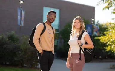 Two students smiling and standing outside on a sunny morning wearing backpacks.