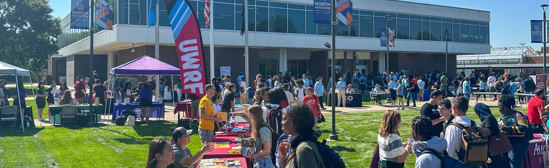 a large group of students outside on a sunny day standing by tables talking to people at the tables. 