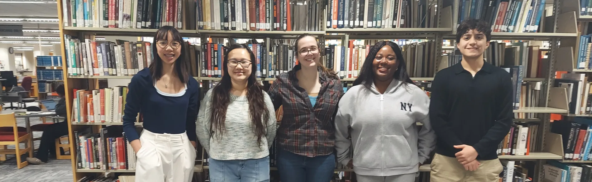 four students and one instructors standing in the library smiling in front of a book shelf. 