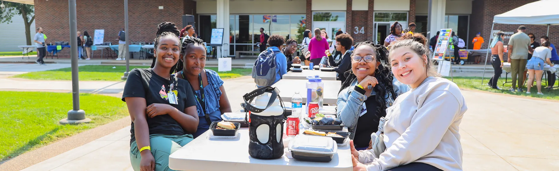 Five students sitting at a table outside smiling on a sunny day with students and a building in the background. 