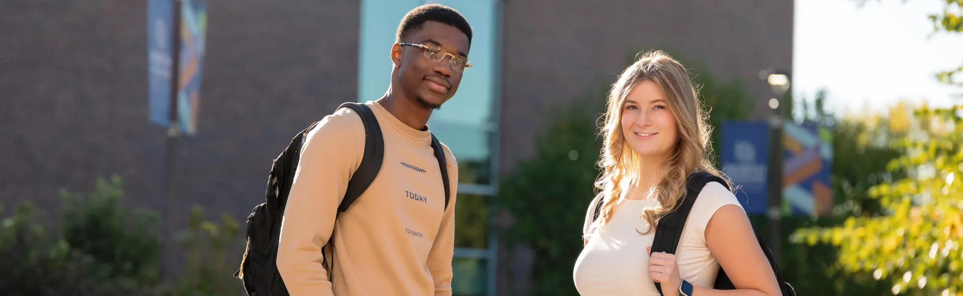 Two students smiling and standing outside on a sunny morning wearing backpacks.