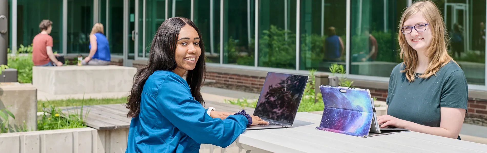 two students sitting at a table with laptops outside on a sunny day. 