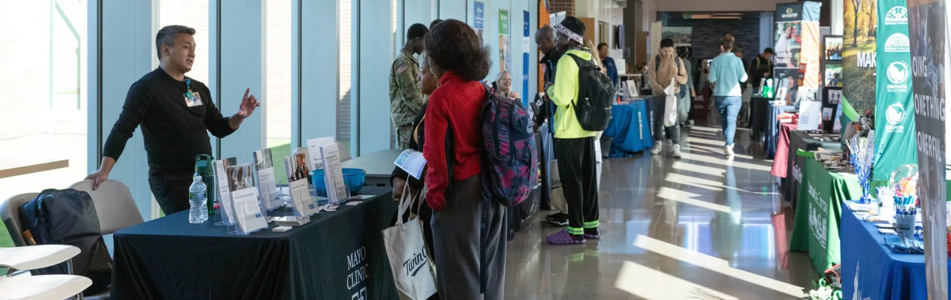 this is a photo of a hallway on our campus during a career fair event. Tables line the walls and students eagerly talk to employers and recruitment staff. 