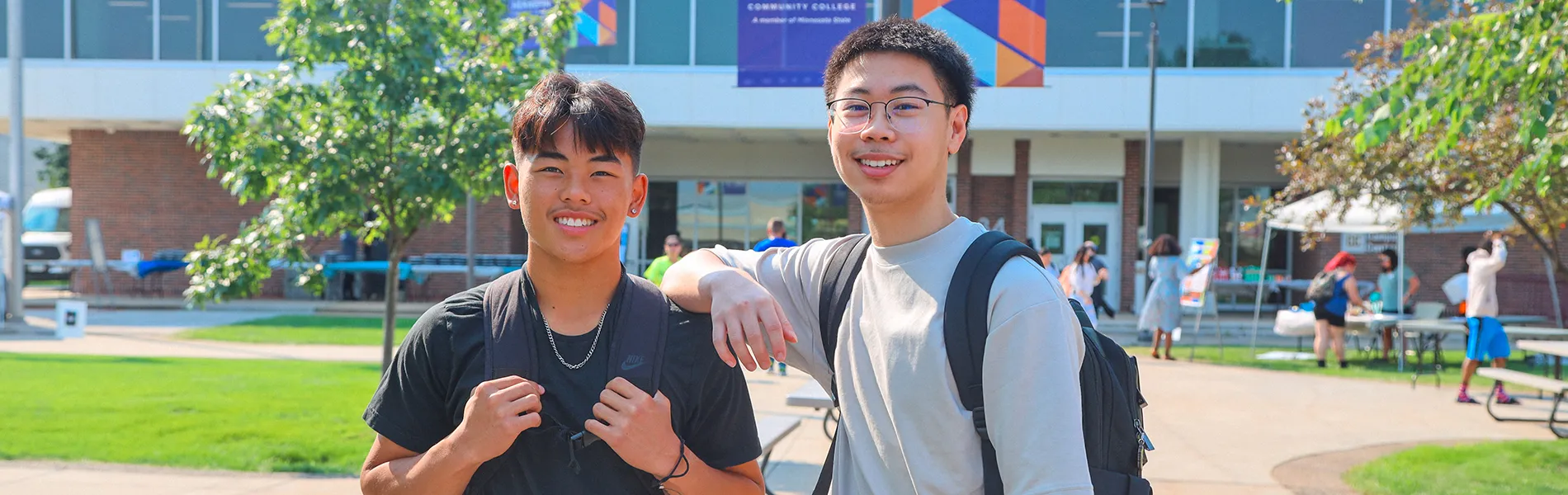two students standing outside smiling on sunny day with college buildings and blue nhcc banners in the background.