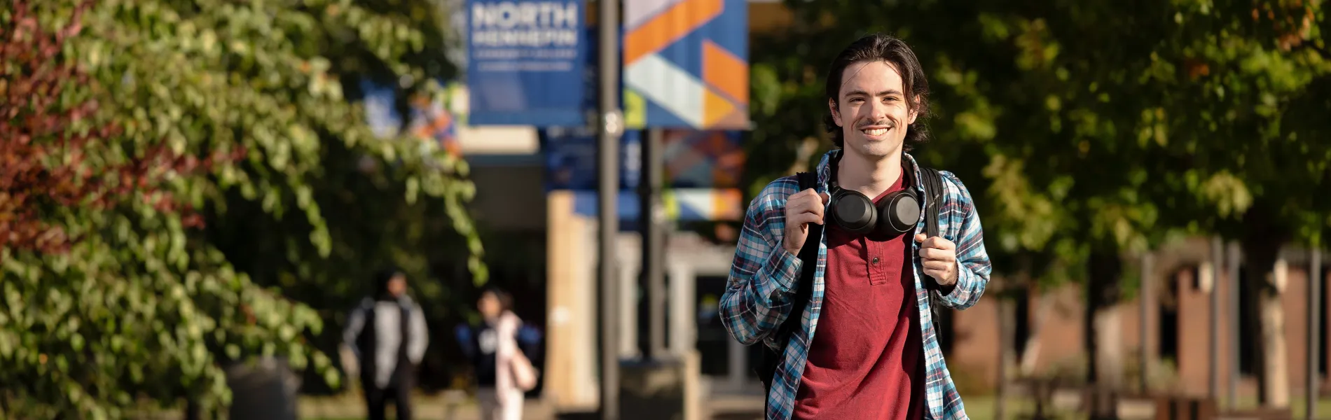 man standing outside smiling wearing a backpack with trees in the background and post with banner that has an NHCC logo on it.