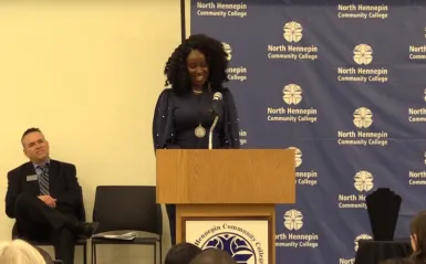 woman presenting on a stage in front of a podium man sitting behind her