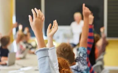 students raising their hands with a teacher in the background 