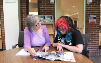 NHCC Workforce Innovation and Experiential Learning Center Director, Karen Philbin, sits with a female student at a table in the library looking at a career magazine together