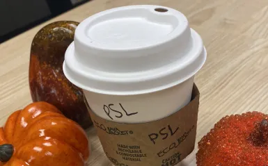 a white, paper coffee cup, with "PSL" written on it in Sharpie marker, next to two mini orange pumpkins 