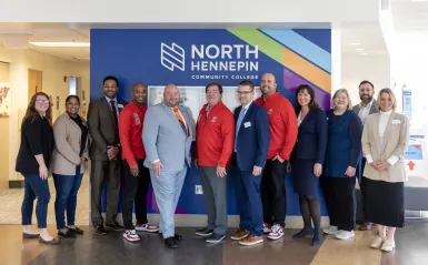 a group photo of staff and administrators from North Hennepin and St. Cloud State University posing for a photo in front of the NHCC sign in the ES building. 