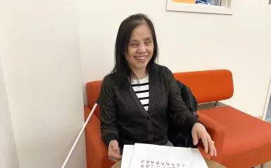 This is a photo of NHCC Student, May Spooner, smiling while sitting in the library on a comfortable chair with her cane at her side and a worksheet of the braille alphabet on the table in front of her along with a braille worksheet on Chapter 4 of the Harriet Tubman textbook she’s reading for her EAP class. 