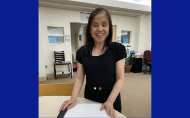 This is a photo of NHCC student, May in the library on-campus, outside of the Access Services offices, standing and smiling beside a table, hands on her newly printed braille textbook. 