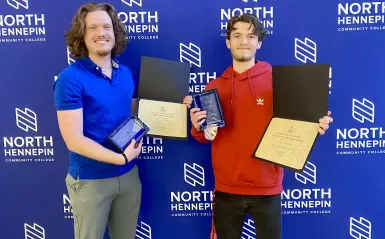 This is a photo of NHCC students and friends, Owen and Travis standing in front of an NHCC logo "step and repeat" backdrop smiling, both holding their certificates and awards, which are plaques made of glass, after being recognized at the Annual Student Life Awards Banquet. 