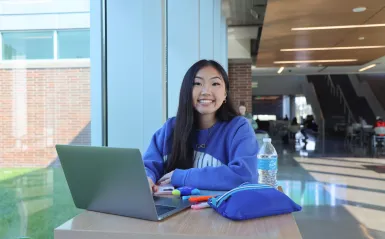 This is a candid photo of a female student smiling for the camera as she looks up from studying at a table in the BHCC hallway. The photo was taken in 2022.