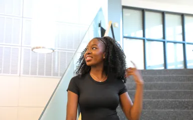 This is a photo of NHCC student and commencement speaker, Oluwaseun Sule, standing on the staircase in the CBT building, smiling and looking off to the side, while she lightly brushes her hand on her beautiful curly, black hair. 