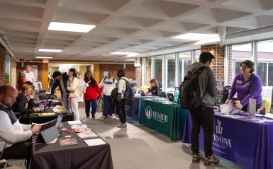This is a photo of students walking through the ES and LRC hallway mingling with representatives from four-year colleges who are sitting there at tables in the hall to tell students about the transfer process. 