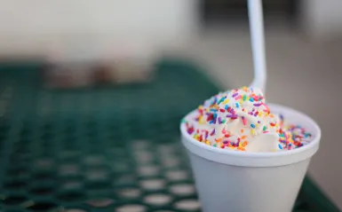 this is a photo of a cup of vanilla ice cream with rainbow sprinkles and a spoon sitting on a picnic table. 