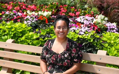 this is a photo of former NHCC student, Estefanny Zelaya, sitting outside on a bench, smiling, with colorful flowers surrounding her. 