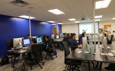 this is a photo of a graphic design classroom in NHCC's Fine Arts Center. Students are working at the iMac desktop computer stations, sitting in comfortable chairs, the teacher is pointing at a graphic on the screen at the front of the room, the wall is bright, dark blue and the vinyl floor is updated. 