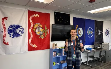 this is a photo of NHCC staff, Dylan Rowinski holding up NHCC's student veteran patches and smiling in our Veterans Resource Center on campus. 