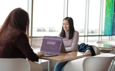 this is a photo of two female students studying at a table in the BHCC building, near a window, with their laptops on the table. One girl is smiling and looking at the camera and you can only see the back of the other girls head. 