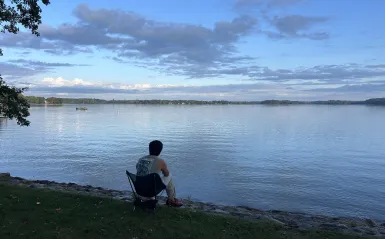this is a photo of a person sitting by a lake with their back to the camera fishing. The sky and the lake are blue. 