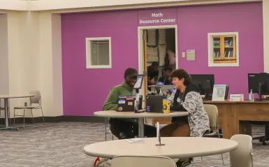 this is a photo of an NHCC tutoring employee sitting at a table working with a student. They are both looking at their open laptops, smiling, at a table in NHCC's Tutoring Center. A purple wall is behind them, with a sign that reads, 'Math Resource Center.'