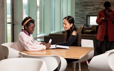 this is a photo of two female NHCC students sitting in chairs at a table by the coffee shop on-campus. The fireplace is in the background, and they are smiling at each other, while looking at papers in a notebook on the table. 