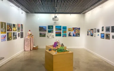 this is a photo of an empty NHCC Art Gallery, with art on the walls and tables, the lights are on and there's a 3D sculpture in the corner from the spring 2025 NHCC Student Art Show. 
