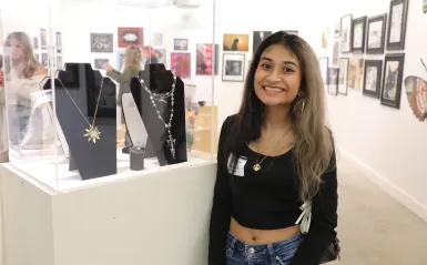 this is a smiling photo of a Maple Grove High School student named Willow Johri taken at the Northwestern High School Art Show Awards Ceremony evening. She is standing in NHCC's Art Gallery, next to a jewelry case with a gold sunshine necklace that she designed in it. 