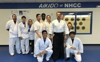 this is a group photo of NHCC instructor, Mark Larson standing with ten of his aikido students in the dojo on-campus, they are all wearing white robes and white pants, smiling for the photo. A sign in the background reads, 'Aikido at NHCC.' 