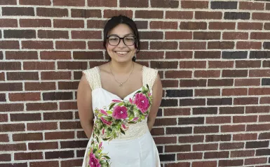 this is a photo of female, NHCC math student, Brenda. She is standing outside, againist a brick wall, wearing a white dress, with handpainted pink roses on it from Mexico. 