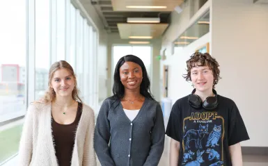 this is a photo of three NHCC students standing side-by-side in the upstairs hallway of the BHCC builidng. Two female students, Anas and Danista stand next to male student, Kenny, posing for a photo and smiling. 