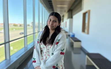 this is a photo of female NHCC student, Reyna Hernandez, standing in the upstairs hall of the BHCC building, smiling, wearing a tank top and a knit cardigan sweater that both have red cherries on them. 