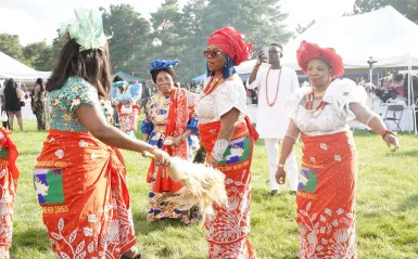 Several attendees wearing vibrant red, white, and multicolored traditional Igbo clothing and headwraps dance in a grassy outdoor festival area during the IgboFest Cultural Celebration. White event tents and other attendees are visible in the background.